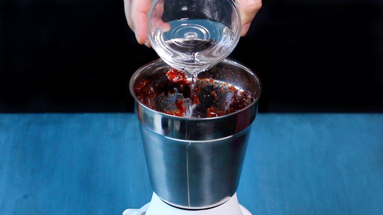 Water being poured from a small glass bowl into a table top metallic grinder to make Date and Tamarind Chutney - Sweet Chutney Recipe