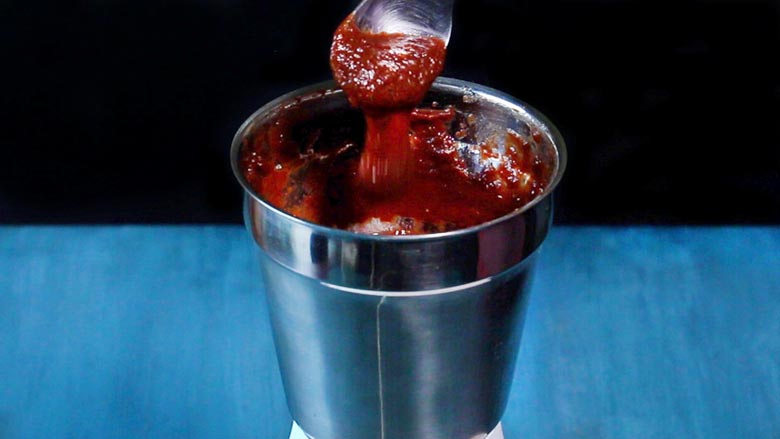 Blended Date and Tamarind Chutney being picked up in a spoon from the metallic grinder to check the consistency of the chutney. On a blue table with a black background. Sweet Chutney Recipe