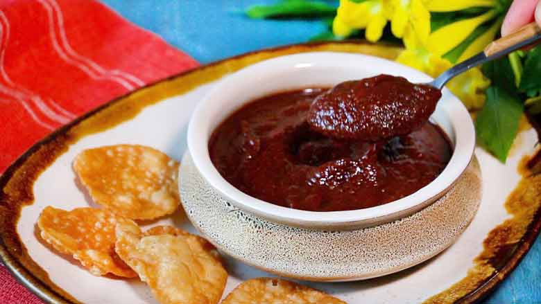 Date and Tamarind Chutney (meetha chutney) being served from a speckled ceramic bowl lying on a blue table with a red cotton cloth and yellow flowers and leaves in the background - Sweet Chutney Recipe