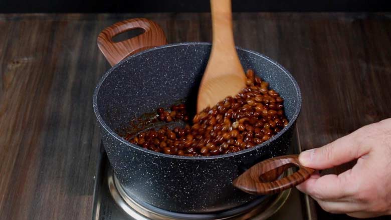 Stirring and cooking kala chana with a wooden spatula in a pan with two large wooden handles on a metallic electric stove on a wooden table - Sukha Kala Chana Masala Recipe