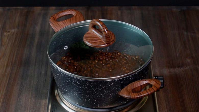 Making Sukha Black Chickpea Masala in a speckled black and white pan with wooden handles covered with a glass lid with a large wooden handle on a metallic stove - Dry Black Chana Masala Recipe