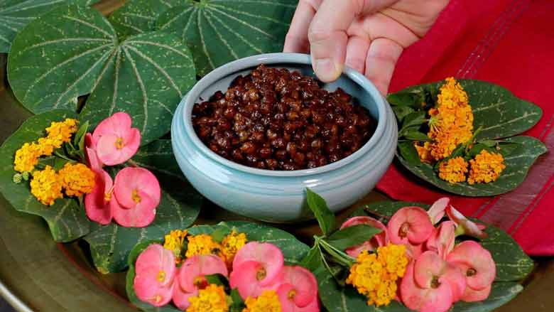 A hand placing an aqua ceramic bowl filled with Sukha Kala Chana Masala on a red fabric surrounded by Indian Orchid leaves and peach, pink, and orange coloured flowers - Sukha Kala Chana Masala Recipe