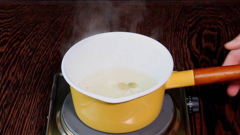 Sugar syrup with green cardamom simmering in a yellow and white saucepan with a wooden handle over an electric stove on a wooden counter - Suji Halwa recipe