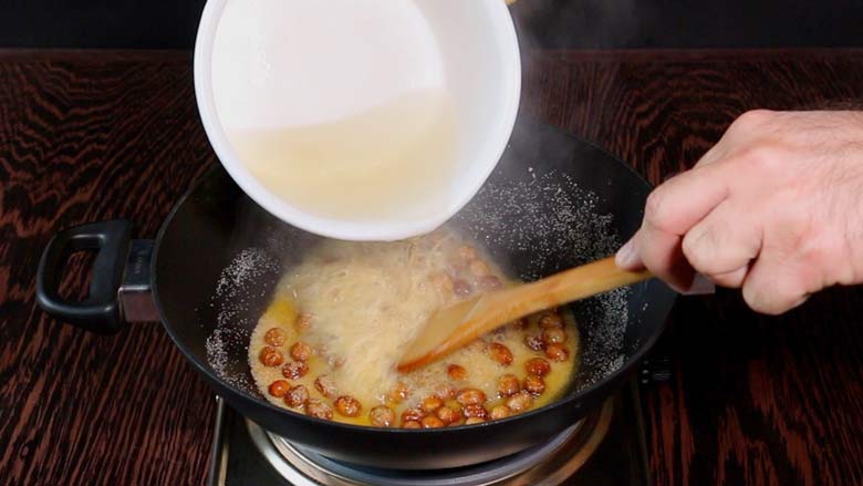 Hot sugar syrup being poured into a black kadhai with the semolina roux - roasted suji, ghee, and kishmish - while a hand stirs with a wooden spatula. Steam is seen rising - Suji Halwa recipe