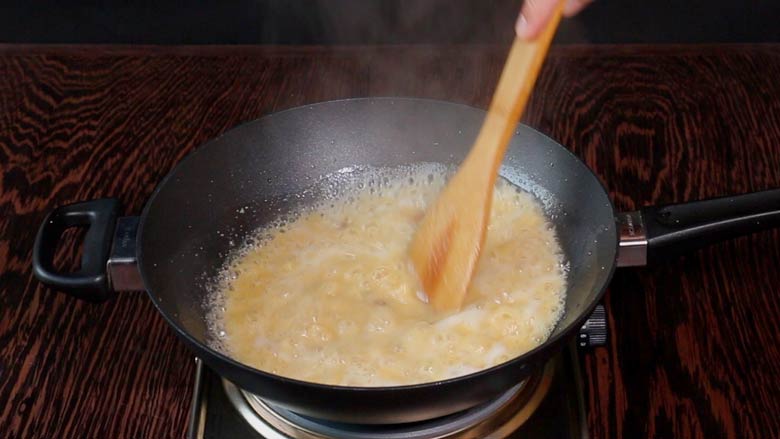 Suji, ghee, and hot sugar syrup bubble altogether in a large black kadhai on a metallic electric stove on a wooden counter while a wooden spatula is seen stirring the halwa - Suji Halwa recipe