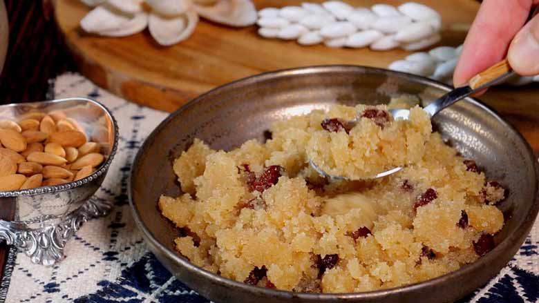 Suji ka Halwa being served using a spoon from a wide bronze coloured ceramic bowl over a blue and white woven table mat. A silver bowl with badam and a wooden cutting board with shell flowers in the background - Suji Halwa recipe
