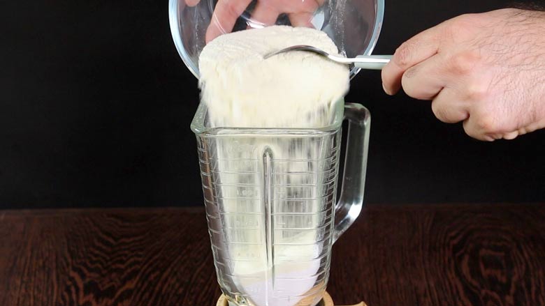Milk powder being tipped in to a blender from a glass bowl with the help of a spoon with a hand visible - Homemade Condensed Milk Recipe