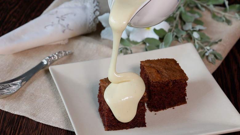 Thick homemade condensed milk being poured over a piece of cake from a white gravy boat lying next to another piece of chocolate cake on a white platter on an ecru linen cloth with a silver spoon and white flowers in the background - Homemade Condensed Milk Recipe