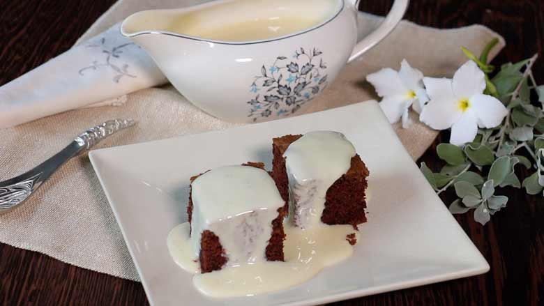 Thick homemade condensed milk drenching two pieces of chocolate chiffon cake on a white platter on an ecru linen cloth with a silver spoon, white flowers and a white floral printed gravy boat full of condensed milk in the background - Easy Condensed Milk Recipe