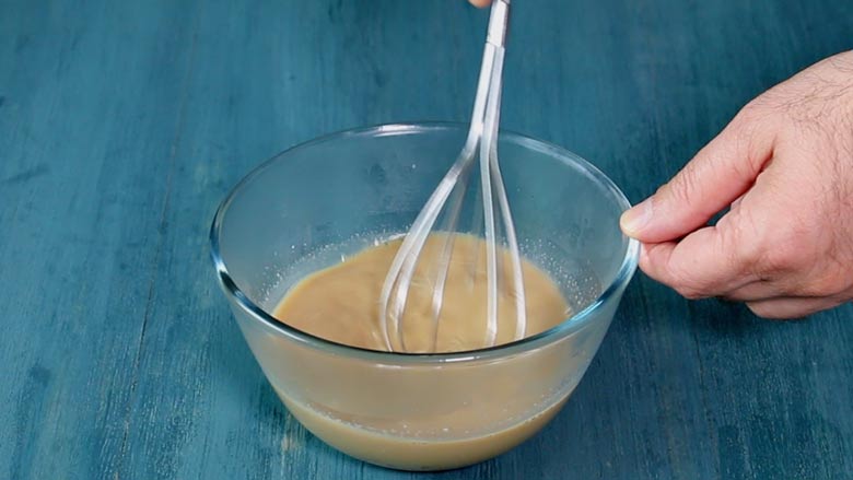 Jaggery syrup in a glass bowl on a blue wooden table being whisked with a balloon whisk - Gulgule Recipe