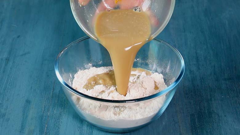 Jaggery syrup in a glass bowl being poured into dry batter ingredients in a larger glass bowl on a blue painted wooden surface - Gulgule Recipe