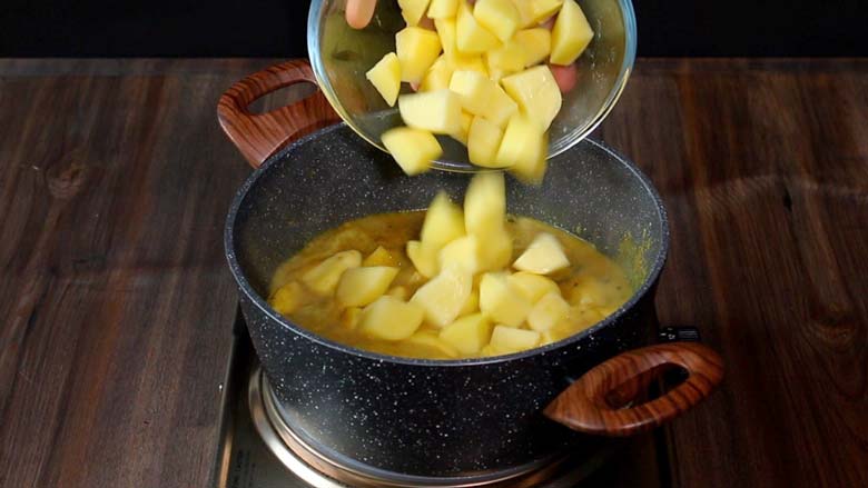 Adding potatoes to the curry cooking in a grey speckled pan with wooden handles on a metal stove while preparing Potato Mappas - Potato Curry Kerala Style