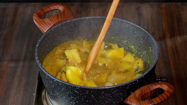 Bringing the potato curry to a boil and stirring with a wooden spatula in a black and white speckled pan on a metal stove on a wooden table -  Potato Mappas Recipe