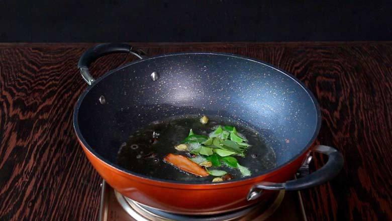 Whole spices and curry leaves being fried in ghee in a large kadhai shaped orange non-stick pan with two handles on a metallic electric stove on a wooden table - Keema Curry Recipe Konkani Style