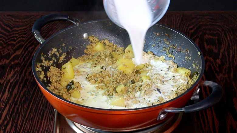 Thick coconut milk being poured in the minced meat curry to simmer briefly in an orange non stick pan on a metallic stove on a wooden table - Keema Curry Recipe Konkani Style