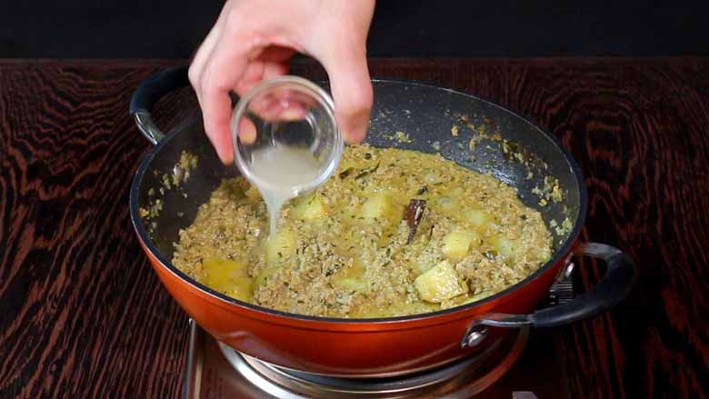 Lime juice being poured from a small glass bowl onto the keema curry in an orange non stick pan on a metallic stove on a wooden table to finish off the dish - Keema Curry Recipe Konkani Style