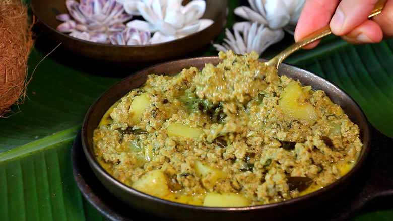 Keema curry with potatoes being served with a bronze coloured spoon from a shallow ceramic bowl lying on a banana leaf with a coconut and some shell flowers in a bowl showing in the background - Keema Curry Recipe Konkani Style