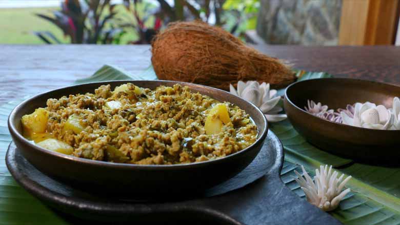 Konkani keema curry plated in a wide ceramic bowl lying on another ceramic platter on a banana leaf with a coconut, shell flowers, and plants in the background - Keema Curry recipe Konkani Style
