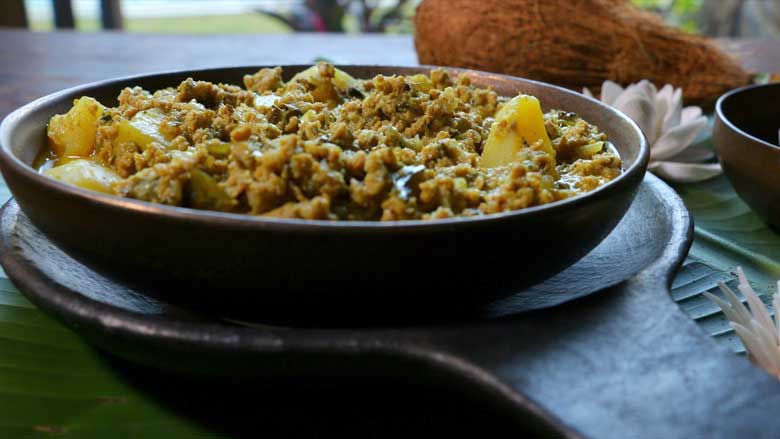 Konkani keema curry plated in a wide ceramic bowl lying on another platter with a single long ceramic handle on a banana leaf with a coconut, shell flowers, and a garden in the background - Keema Curry recipe Konkani Style