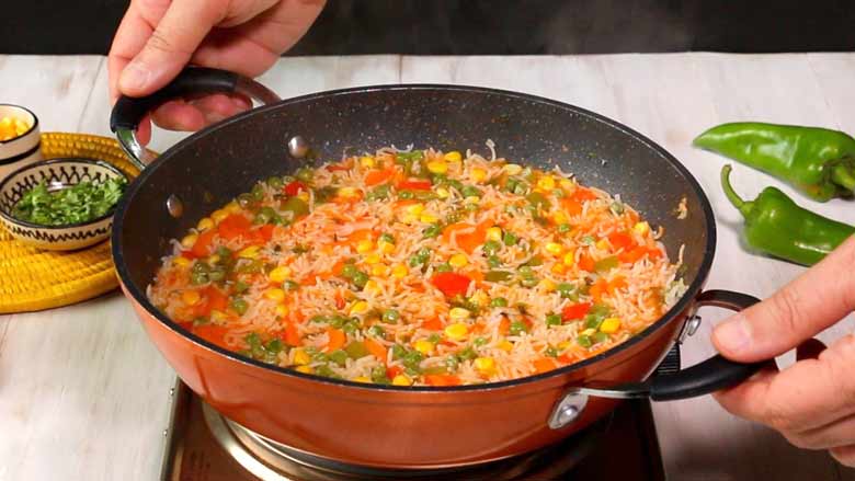 Hands are seen taking cooked and ready Mexican rice off the heat in a black and orange non stick pan. Two large green chillies and parsley and corn in small bowls lie in the background - Arroz a la Mexicana recipe