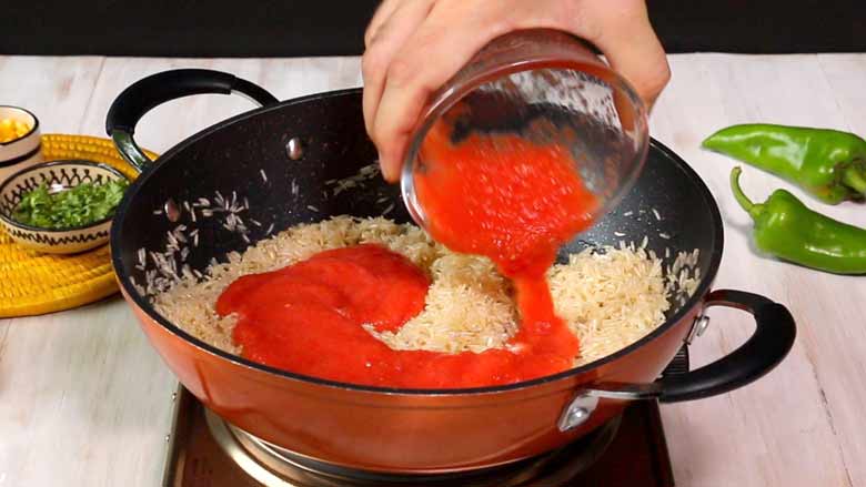 Tomato garlic puree is being poured from a glass bowl over rice toasting in an orange and black non stock pan with two handles. Two large green chillies and parsley and corn in small bowls lie in the background - Arroz a la Mexicana recipe