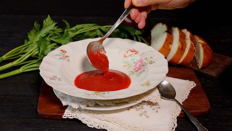 A hand is seen ladling ruby red borscht soup into an antique English floral bone china soup bowl lying on another plate with lace napkins underneath. Celery and bread lie in the background - Borscht Soup Recipe