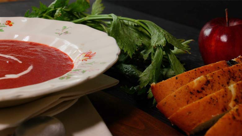 Ruby red Russian borscht soup in an antique English floral bone china soup bowl is seen partially on the left side of the image with sliced bread, celery, and an apple are seen on the right side in a country setting - Borscht Soup Recipe
