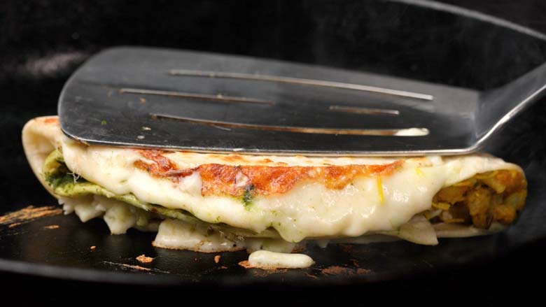 A steel spatula is seen pressing down on a tortilla wrap being grilled in a black cast iron pan with melting cheese oozing out - Cheesy Tortilla Wrap Recipe