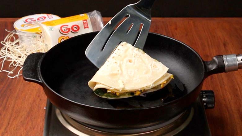 A steel spatula places a cheese tortilla wrap into a heavy cast iron pan on a stainless steel electric stove over a natural wood counter - Cheesy Tortilla Wrap Recipe
