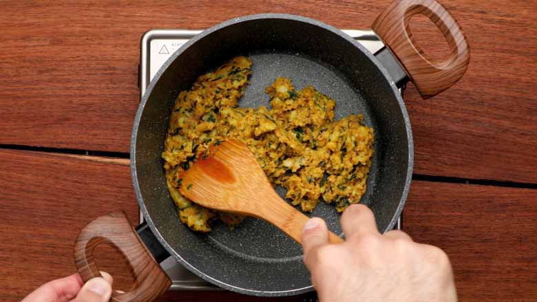 A hand is seen stirring masala potatoes with a wooden spatula being cooked in a grey non stick pan with two large wooden handles over a stainless steel electric stove placed on a natural wooden surface - Cheesy Tortilla Wrap Recipe