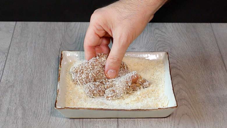 A hand is seen dredging a marinated boneless thigh of chicken in panko breadcrumbs so it is coated well with breadcrumbs in a square white ceramic platter on a grey wooden counter - Chicken =Farcha Recipe