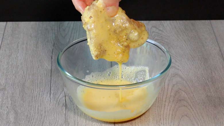 A crumbed thigh of chicken coated with beaten egg is seen held dripping over a glass bowl of beaten egg on a grey wood table top before being fried - Chicken Farcha Recipe