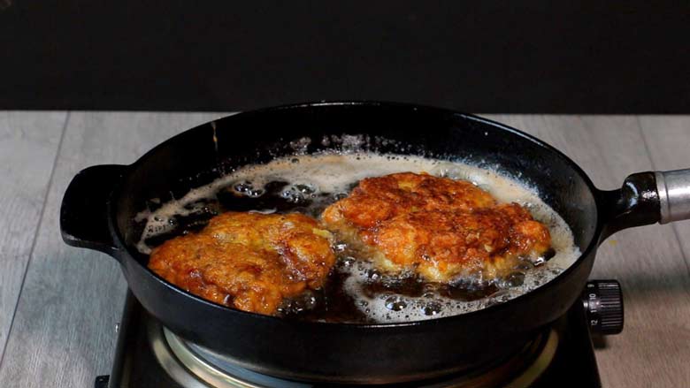 Two pieces of Parsi style crispy fried chicken being fried to golden brown in a cast iron frying pan on a metallic electric stove on a grey wooden counter - Chicken Farcha Recipe
