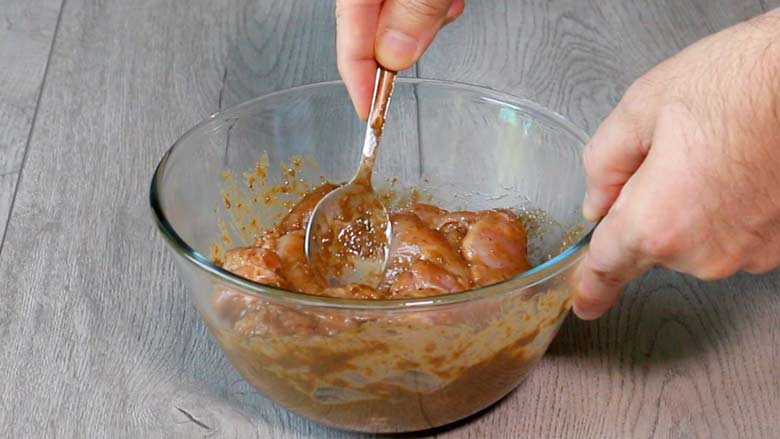 Boneless thighs of chicken in a glass bowl being mixed with a spoon in a marinade made of spices, vinegar, and oil - Chicken Farcha Recipe