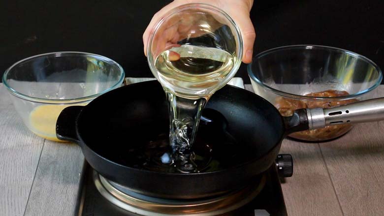 Oil being poured from a glass bowl into a cast iron pan on a metallic electric stove on a grey wooden table with bowls of beaten egg and marinated chicken in the background - Chicken Farcha Recipe | Parsi Style Fried Chicken