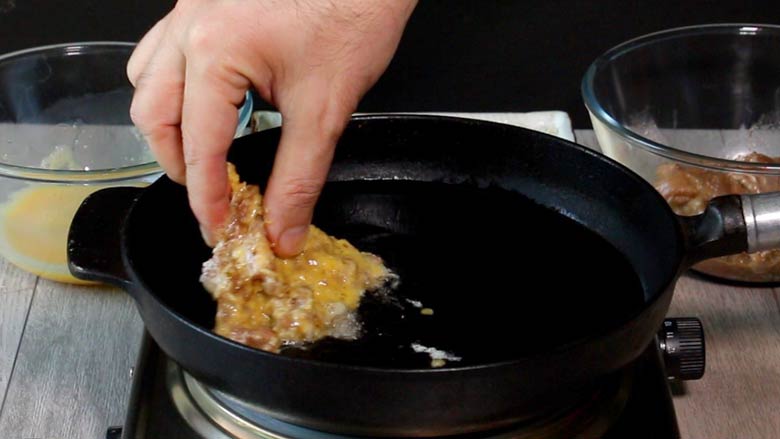 A hand is seen placing a crumbed, egg coated chicken thigh gently in hot oil in a cast iron frying pan with bowls of beaten egg and marinated chicken in the background - Chicken Farcha Recipe | Parsi Style Crispy Fried Chicken