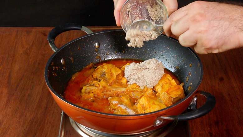 A hand is seen spooning prepared kodi vepudu curry masala paste from a small glass bowl into the Andhra chicken fry cooking in an orange and black nonstick pan with two handles on an electric stove on a wooden countertop - Masala Chicken Fry Recipe