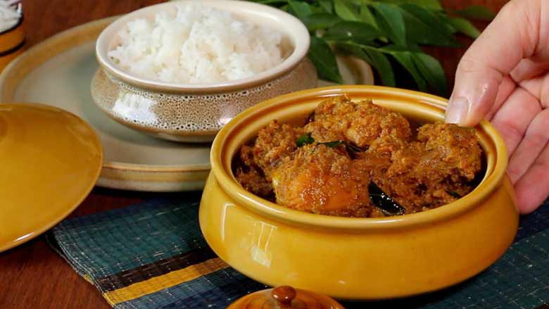 A hand is seen placing a bowl of cooked Andhra chicken fry on an aqua and yellow striped mat on a table with a bowl of rice and fresh curry leaves in the background. The ochre lid of the bowl is seen on the side - Chicken Masala Fry Recipe