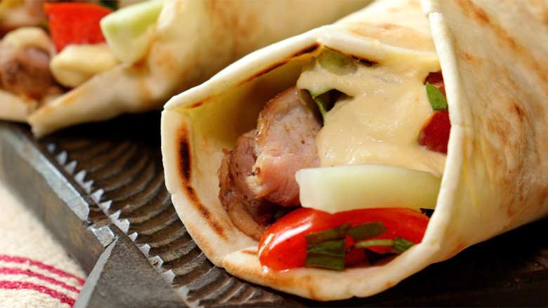 A closeup shot of a chicken shawarma with sliced chicken, tomato, hummus, and pickled cucumber showing in the wrap with another shawarma in the background - Homemade Chicken Shawarma Recipe