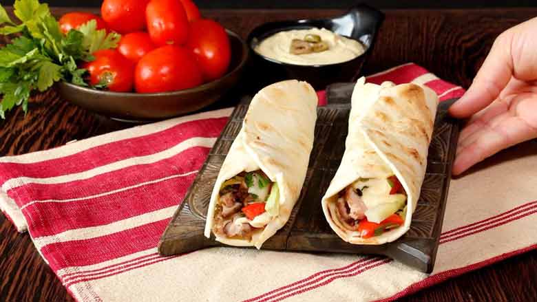 A hand is seen placing a small wooden platter with two chicken shawarma on it onto a woven striped red and cream tablecloth with a bowl of hummus with olives and a larger bowl with whole Roma tomatoes and parsley in the background - Homemade Chicken Shawarma Recipe