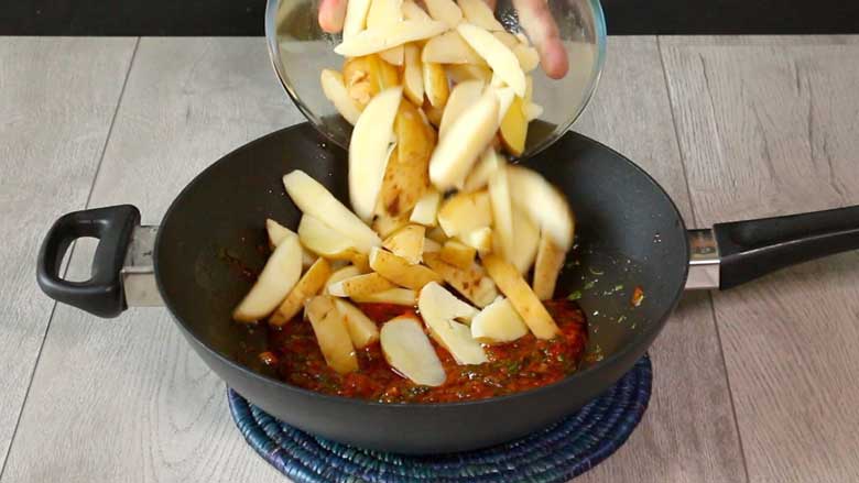 Boiled potato wedges with skin on are being tipped into a kadhai holding the chilli dressing / sauce. The kadhai is on a blue cane mat on a grey countertop - Chilli Potato Salad Recipe
