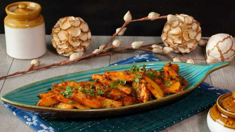 A side shot of chilli potato salad served in an leaf shaped aqua striped ceramic platter on a dark green mat with a printed cloth border with paper flowers and ceramic martbans in the background - Chilli Potato Salad Recipe