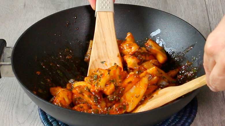 The chilli flecked potato salad is being gently tossed using two wooden spatulas in a black kadhai lying on a blue mat - Chilli Potato Salad Recipe