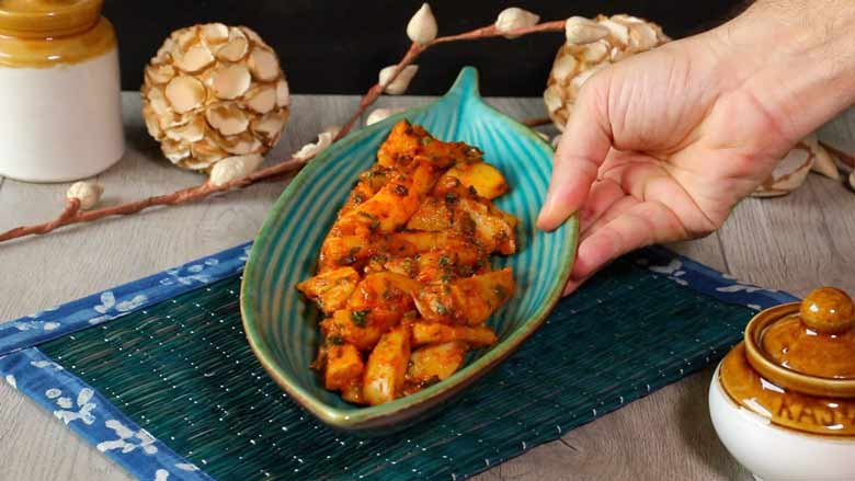A hand is placing an aqua striped ceramic platter shaped like a leaf filled with chilli potato salad on a dark green mat with a printed cloth border with paper flowers and ceramic martbans in the background - Chilli Potato Salad Recipe