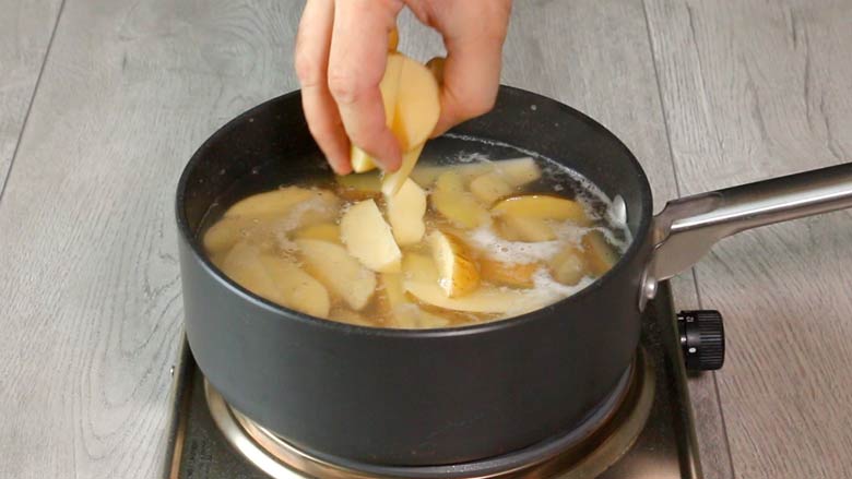 A hand is seen adding potato wedges to boiling water in a dark grey saucepan on a metallic stove on a grey counter top - Chilli Potato Salad recipe