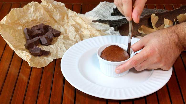 A molten lava cake in a ramekin lies on a white plate while a hand runs a knife around its edge to loosen the cake. The other hand hold the ramekin. Chocolate pieces on a brown parchment paper and dried leaves and bark is see in the background on a slatted wooden counter - Chocolate Lava Cake