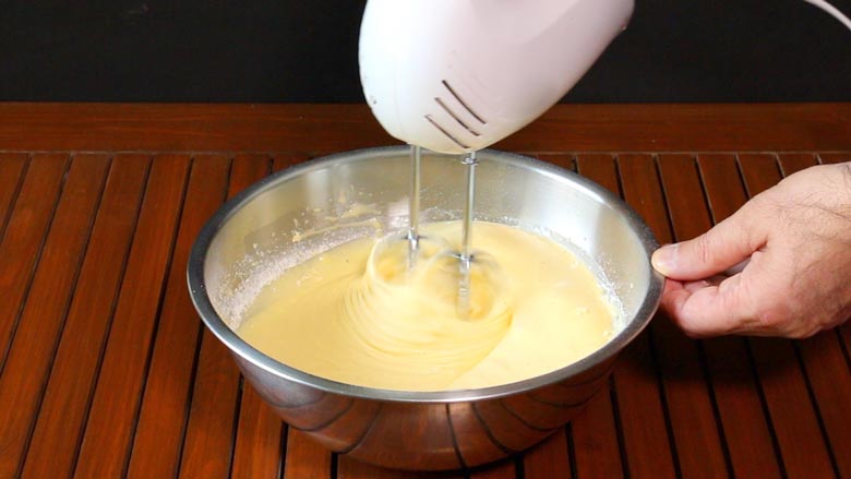 Eggs and sugar are being whisked in a large metallic whisking bowl with a hand held electric whisk while one hand holds the bowl. The bowl lies on a slatted wood table - Chocolate Lava Cake Recipe