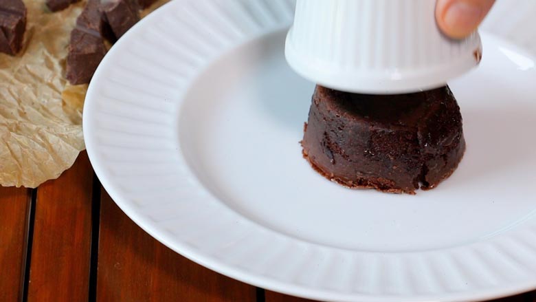 The ramekin is seen being lifted off the flipped molten chocolate cake on a white plate on a brown slatted wood table - Chocolate Lava Cake Recipe
