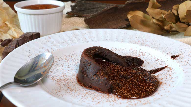 Chocolate lava cake on a white plate oozing melted chocolate has been dusted with cocoa powder. A ramekin with another chocolate lava cake, brown parchment paper with chocolate pieces and dried leaves and bark are seen in the background - Chocolate Lava Cake Recipe