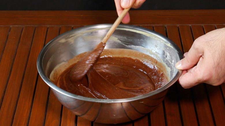 A hand is seen holding a spatula and gently folding the egg and chocolate mixture together in a large metallic mixing bowl on a slatted wooden table to make the batter for molten chocolate cake - Chocolate Lava Cake Recipe